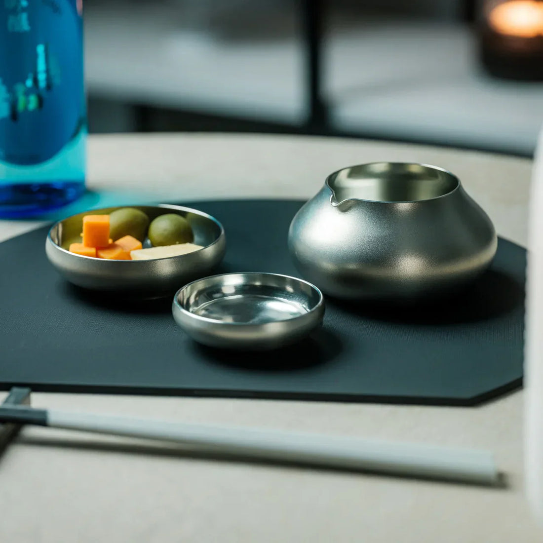 特寫現代餐桌設置，展示精緻的不銹鋼清酒杯和碗，包括一個酒壺和一個裝有奶酪塊和橄欖的小碟，放置在黑色墊子上。背景中的藍色瓶子和蠟燭營造出溫馨的氛圍，非常適合日式用餐體驗。